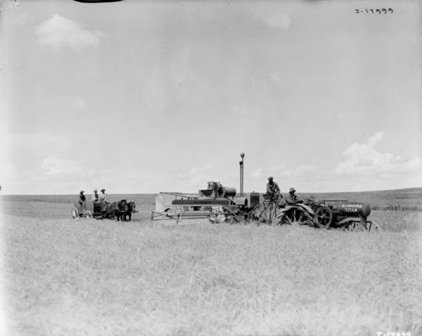 View across field towards two men using a Titan tractor to pull a harvester thresher in a field. Three men sit in a horse-drawn wagon behind on the left. Part of the push binder Titan trials.