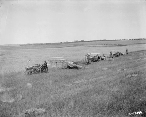 Elevated view of three men on Titan tractors pulling three binders. A  man is standing on the back of each push binder. American Flags are on the front of each tractor. Part of the push binder Titan trial. There are buildings in the far background.