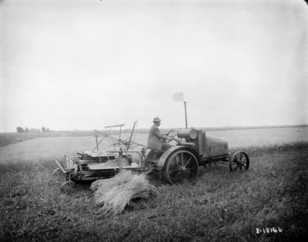 Right side profile view of a man using a kerosene tractor to pull a binder in a field. There is a small U.S. Flag flying from the tractor exhaust pipe.