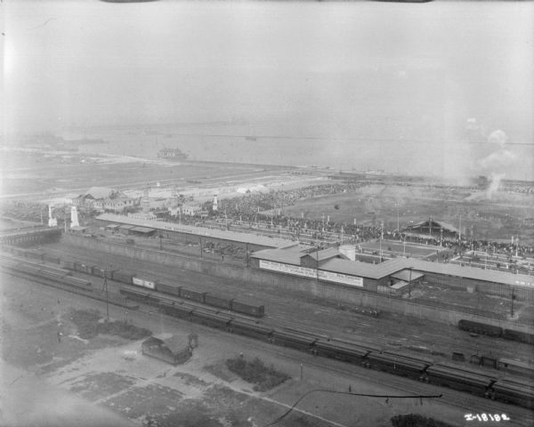 Aerial view looking over railroad cars on railroad tracks towards an encampment. Signs on a building at the encampment read: "War Exposition," and "Three Miles of Trenches! Actual Battle." A large crowd surrounds a field near the buildings and rows of tents. In the background is the shoreline of a lake.