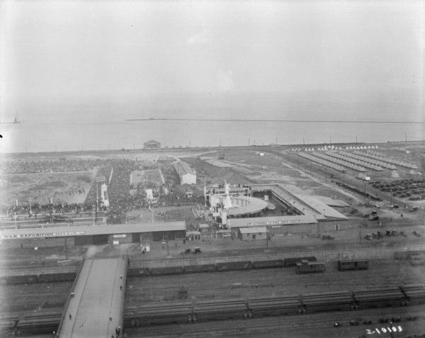 Aerial view looking over railroad cars on railroad tracks towards an encampment. Sign on a building at the encampment reads: "War Exposition." A large crowd surrounds a field near the buildings and rows of tents. In the background is the shoreline of a lake.
