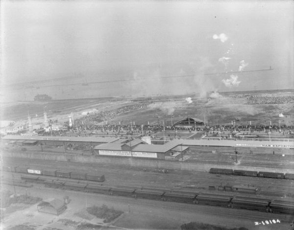 Aerial view looking over railroad cars on railroad tracks towards an encampment. Signs on a building at the encampment read: "War Exposition," and "Three Miles of Trenches! Actual Battle." A large crowd surrounds a field near the buildings and rows of tents. In the background is the shoreline of a lake or ocean.