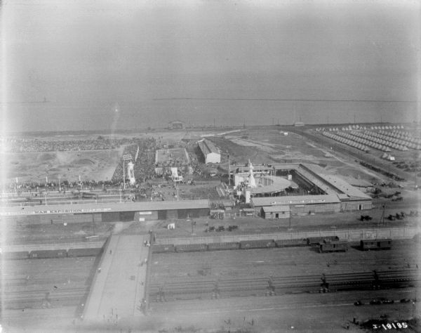 Aerial view looking over railroad cars on railroad tracks towards an encampment. Signs on a building at the encampment read: "War Exposition," and "Three Miles of Trenches! Actual Battle." A large crowd surrounds a field near the buildings and rows of tents. In the background is the shoreline of a lake or ocean.