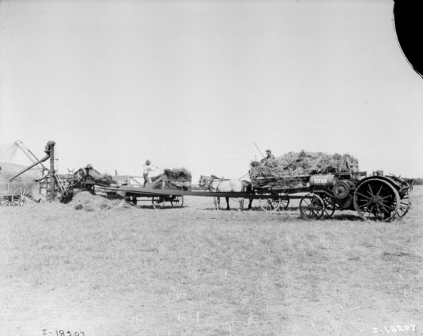 Titan tractor belt-driving a corn separator in a field. Men are working with horse-drawn wagons near the tractor.