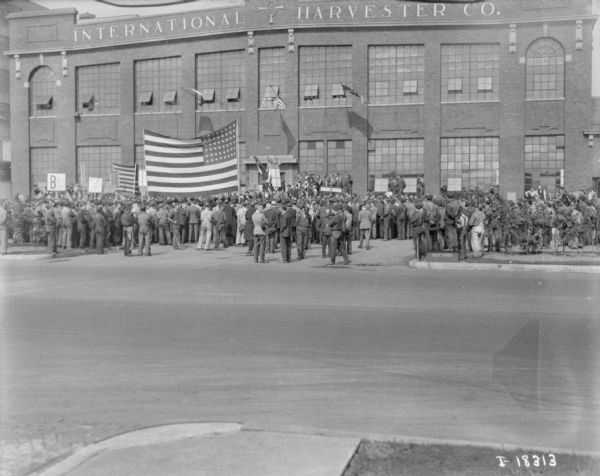 View across street towards a large group of people gathered in front of the International Harvester Co. plant. Some of the people in the crowd are holding up a large U.S. flag. Near the entrance is a group of people who appear to be on a stage. Perhaps the end of WWI.
