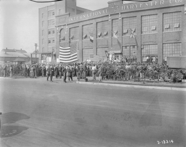 View across street towards a large group of people gathered in front of the International Harvester Co. plant. There are two or three large U.S. flags on display. Near the entrance is a group of people who appear to be on a stage. Perhaps the end of WWI.