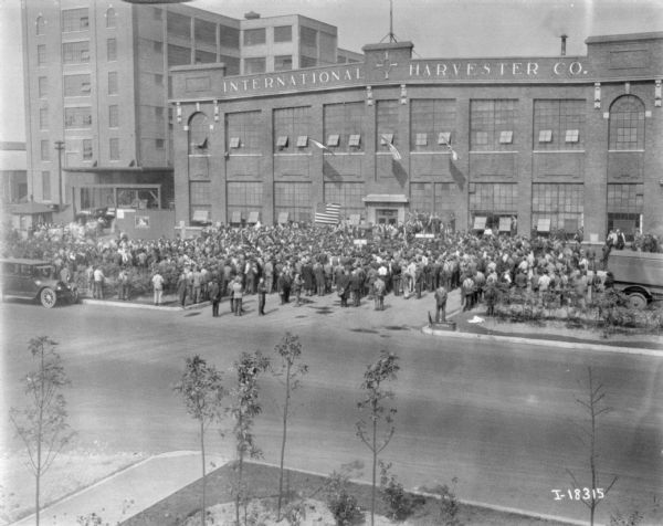 Elevated view across street towards a large group of people gathered in front of the International Harvester Co. plant. Near the entrance is a large U.S. flag and a group of people who appear to be on a stage. Perhaps the end of WWI.