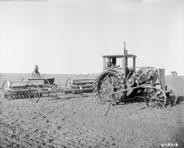 Three-quarter view from right front of a man driving a tractor pulling seeders. Another man is sitting on a seeder in the center directly behind the tractor.