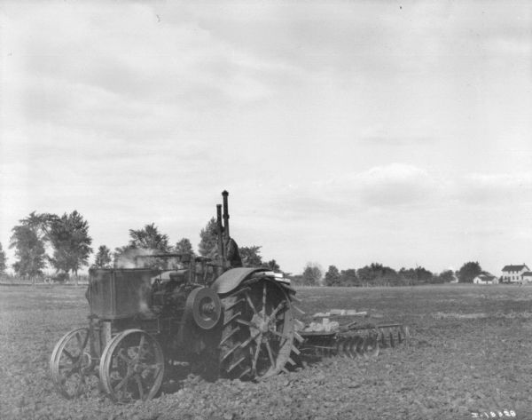 Left side view of man driving tractor pulling a disk harrows. Farm buildings are in the far background on the right.