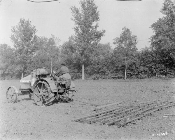 Three-quarter view from left rear of a woman driving an International tractor pulling a peg tooth harrow in a field. The harrow is attached with a chain.