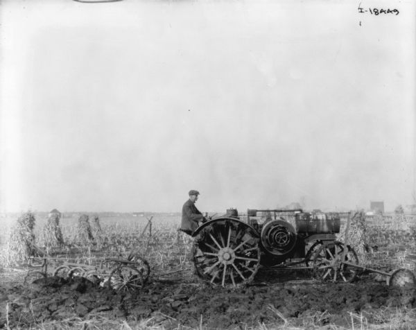 Right side view of a man using a Titan tractor to plow a field. Shocks of grain are in the field on the left. Buildings are in the far distance.