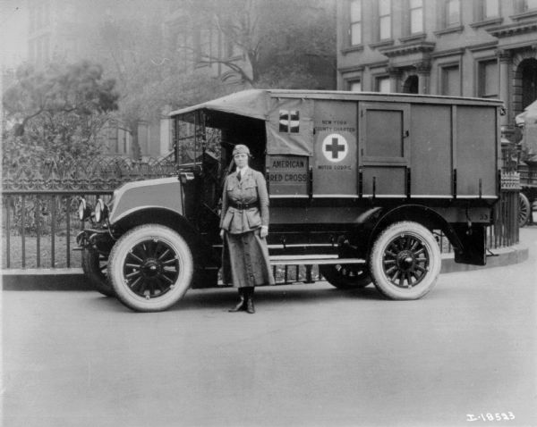 A woman in a uniform posing standing in front of an American Red Cross ambulance which is parked along a fence. Brownstone buildings are in the background. A sign on the truck reads: "New York County Chapter Motor Corps."