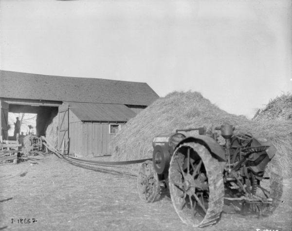 View of a tractor belt-driving a thresher which is at the entrance to a barn. Men are working inside the barn. There are large haystacks behind the tractor.
