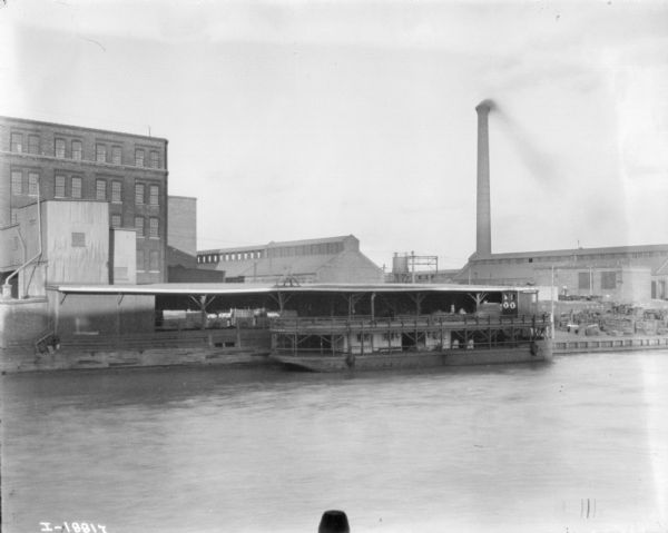 View across water towards factory buildings on the opposite shoreline. A two-decker boat with the name "Harvester" is moored at the dock. A large smokestack is in the background.