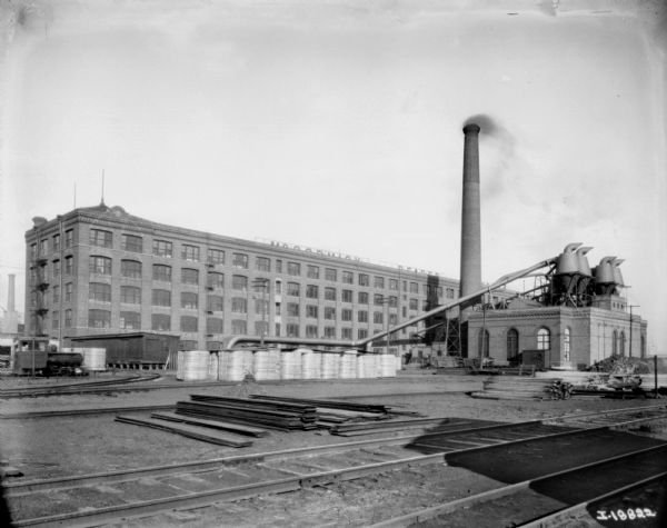 View of the railroad yard at McCormick Works. Railroad tracks are in the foreground, and there is a locomotive near a railroad track turnaround. There is a large brick factory building is on the other side of the yard. On the right is a smokestack, and a smaller, brick building connected to the large building with large pipes.