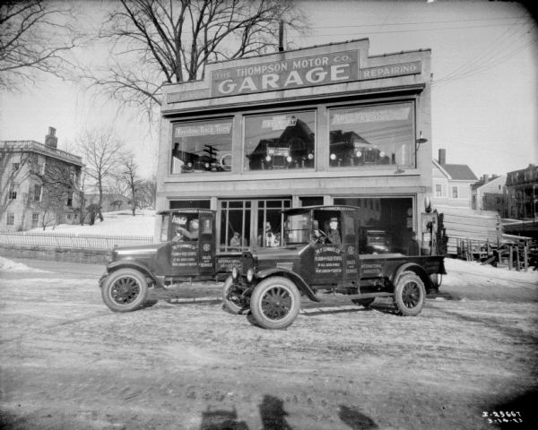 View of two men posing while sitting in the driver's seats of two trucks parked in front of a dealership. The doors of the trucks have painted signs that read: "The P. Schwartz Co." Behind the show windows of the shop, men are standing and looking out at the trucks. Another man is standing near a gas pump on the right. There are large, second-story show windows, with automobiles parked and on display inside.
