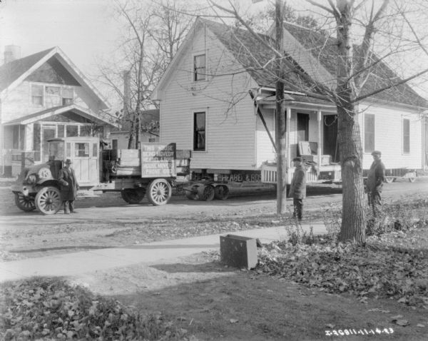 Three men standing near a house on a trailer attached to a truck. A sign on the truck reads: "This house being moved by Heabel & Rose House Movers Phone 1080."