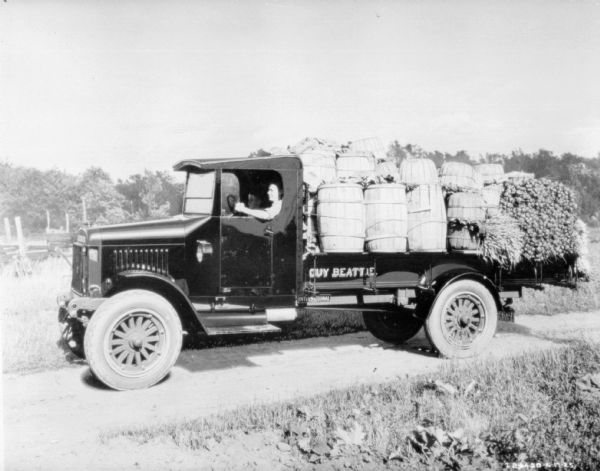 Left side view of a man driving a truck on a road. The bed of the truck is loaded with overflowing barrels and bushel baskets. A young boy is sitting in the passenger seat.