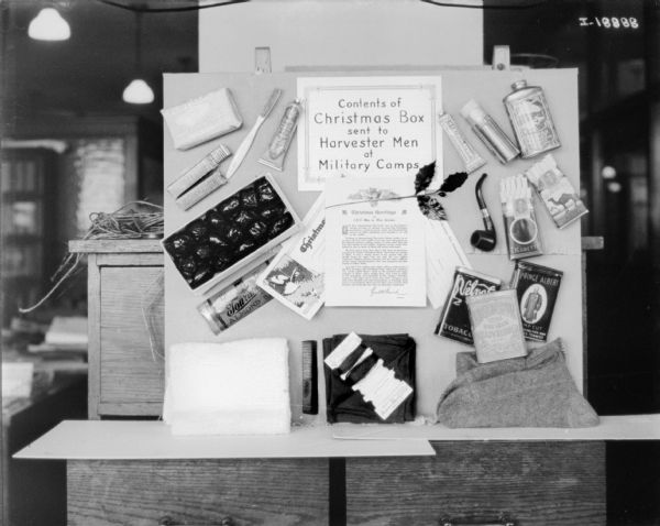 Contents of X-mas box sent to Harvester men at military camps displayed on a wall at McCormick Works. Some of the contents include candy, gum, toothpaste, toothbrush, soap, a pipe, two packs of cigarettes, a comb, and a pair of socks.