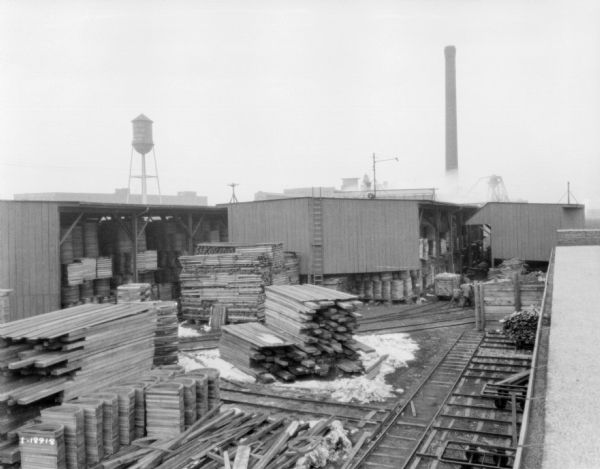 Elevated view from roof of railroad tracks and stacks of lumber in a yard near a factory. Two men are working in the yard in the background on the right. There is a water tower and a smokestack in the distance.