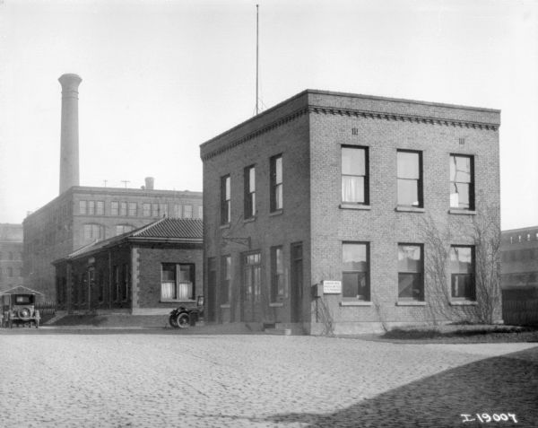 Office building in plant yard. In the background are other brick buildings, automobiles, and a smokestack.