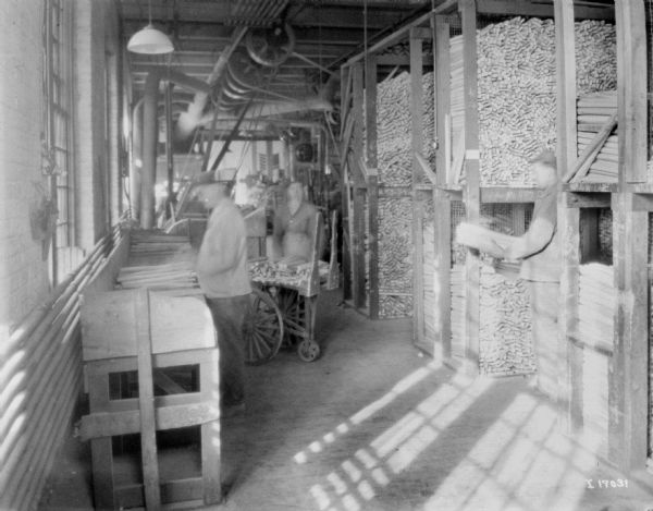 Three people are working in a storage area of a factory. Sun is streaming in from windows on the left. One man on the right holding a bundle of wood while standing among tall shelves holding wood pieces. Two other people are in the center, one working at a table, and another standing behind a cart. Belt-driven machinery is in the background.