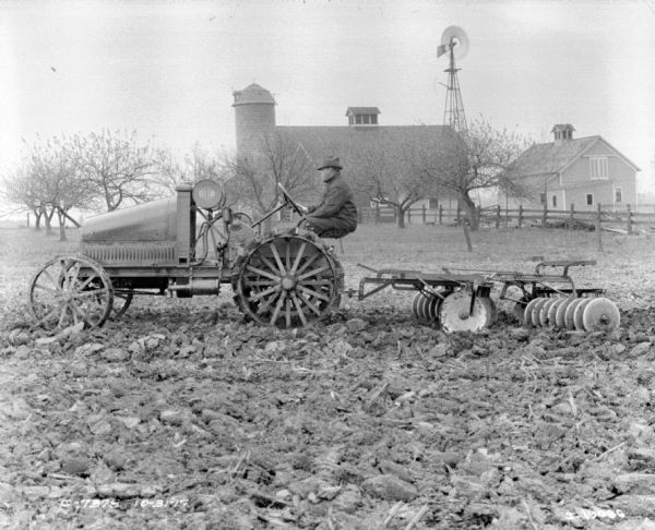 Left side profile view of a man driving a tractor pulling a disk harrow in a field. In the background is a silo, barn, windmill and farmhouse.