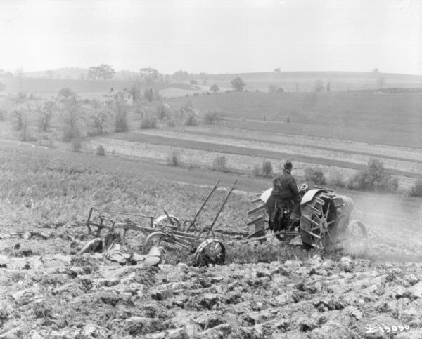 View down hill towards a man plowing with a tractor in a field. Below is a farmhouse among trees.