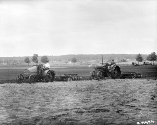 View across field towards two men using tractor to pull disk harrows. In the background another tractor and two automobiles are parked.
