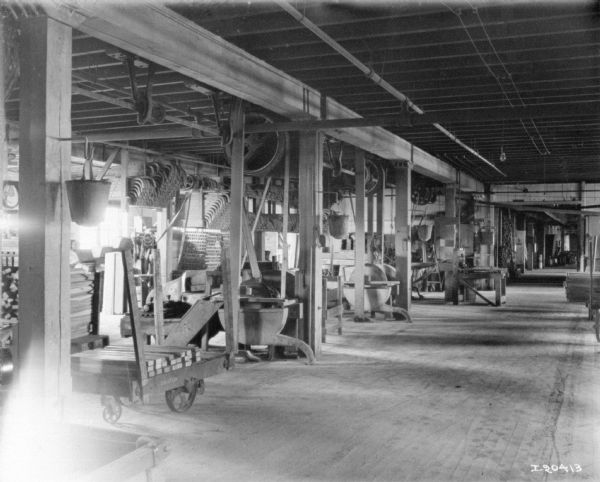 Machinery in the center of the room is belt-driven from large wheels attached to the ceiling. There are parts on a cart nearby, and parts are stacked in the background.