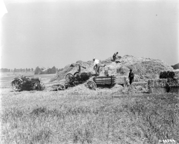 A Titan tractor is belt-driving a hay press. Men are working near a large pile of hay.