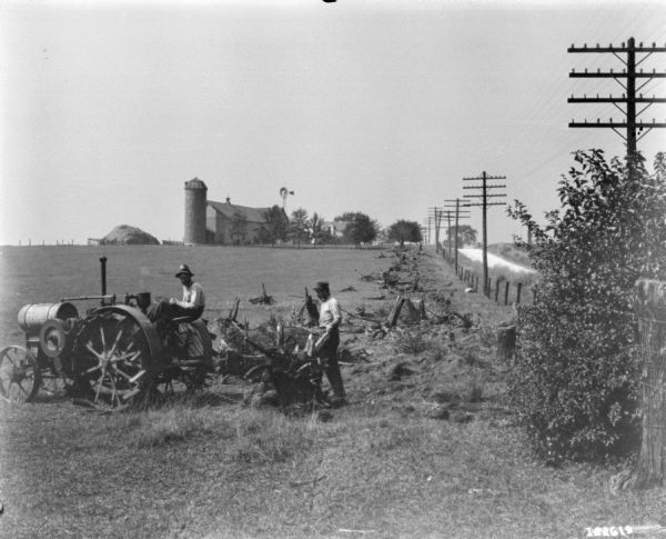 Two men are pulling tree stumps in a field with a Titan tractor. A silo and farm buildings are in the background.