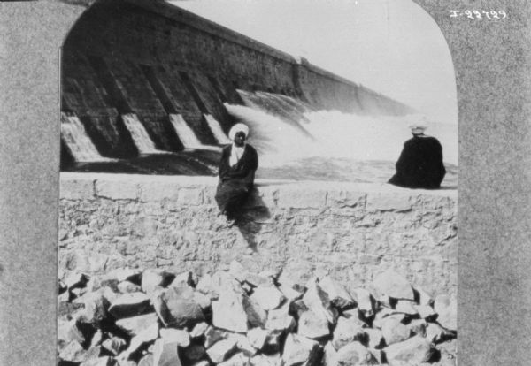 Two men wearing turbans are sitting on a stone wall. There is a hydroelectric dam in the background.