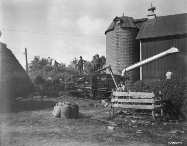 Men are working around a tractor-powered threshing operation. A silo and a barn are in the background.