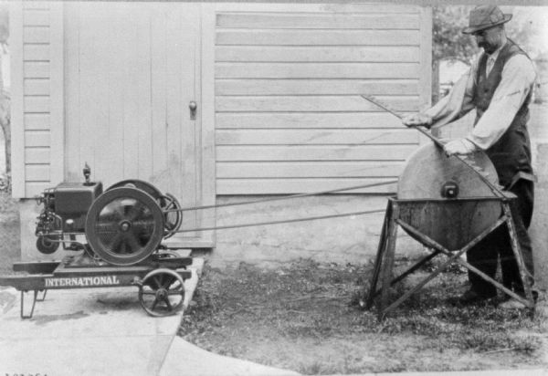 International engine powering a blade sharpener. A man standing on the right is holding a blade to be sharpened against the stone.