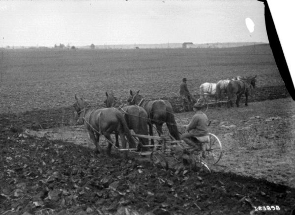 Slightly elevated view of two men, each one driving a team of three mules to pull cultivators in a field in early spring.