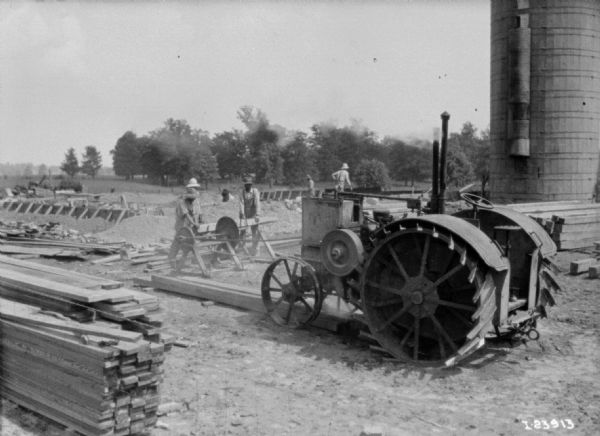 Men using a sawmill powered by a tractor. There is a silo in the background on the right.
