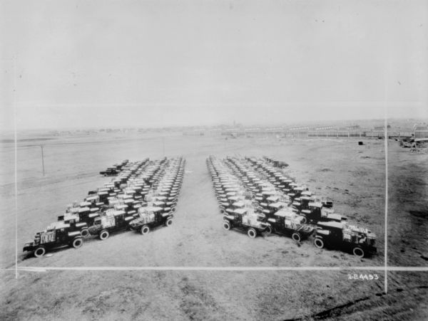 Elevated view of trucks lined up at an angle in two separate, large groups in a field. Equipment is loaded in the back of each truck bed. Factory buildings are in the far background. 