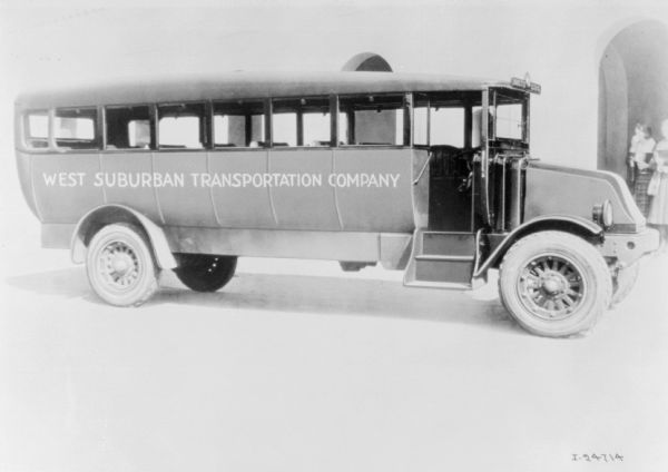 A bus is parked in front of a building. The sign painted on the side of the bus reads: "West Suburban Transportation Company."There are two women standing in an archway on the far right.