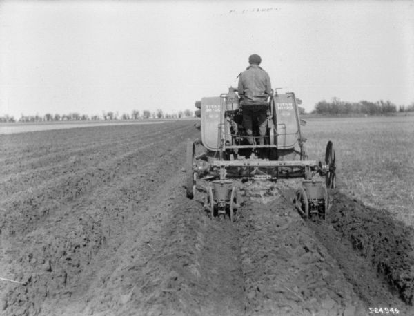 View from rear of a man driving a Titan 10-20 tractor pulling a corn planter in a field.