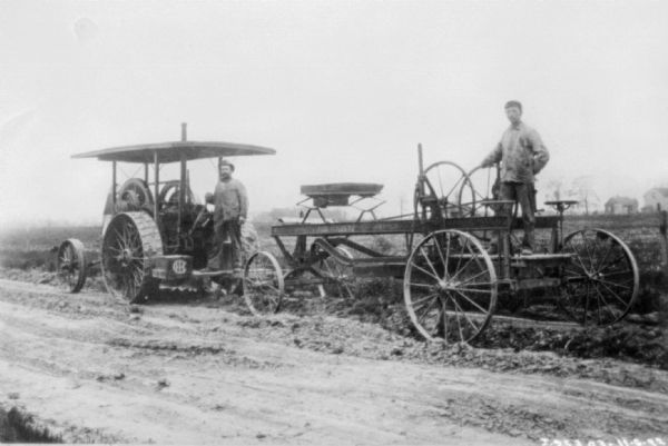Two men in work clothes are standing posing on construction equipment. On the left is a tractor, and on the right a piece of road building equipment. Both are paused on a road. There are buildings in the far background on the right.