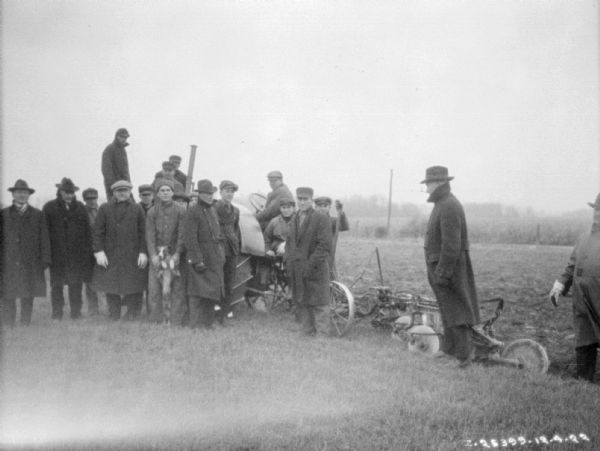 A group of men are posing on an in front of a tractor pulling a plow in a field. One of the men is holding a dog.