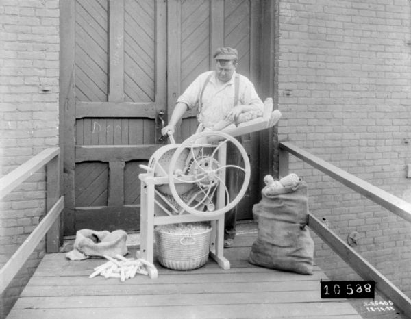 Man shredding corn cobs with a hand-cranked feed mill. He is standing on a wood platform near a the closed double doors to a building. A burlap sack full of dried corn cobs is on the platform on the right. The shelled corn is falling into a metal tub underneath the mill.
