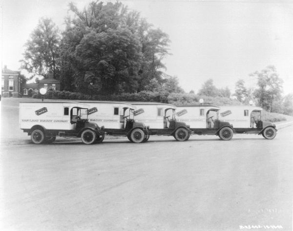 Five delivery trucks parked together at an angle. A large building is in the background. Signs on the trucks are advertisements for "Marbis, Maryland Biscuit Company."