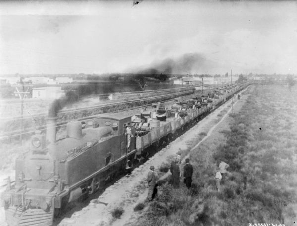 Elevated view of a train with cars loaded with International machines. People are standing near the locomotive on the right.