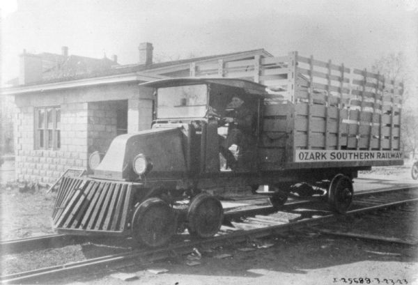 Three-quarter view from front left of man operating a truck adapted to run on railroad tracks. A sign on along the truck bed reads: "Ozark Southern Railway."