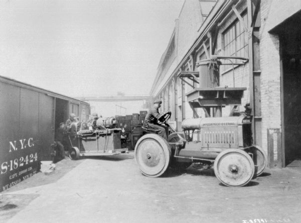 A man driving an industrial McCormick Deering tractor is pulling a sled full of goods from a train car. Three men are helping to push it onto the loading dock. On the right is a large open door to an industrial building.