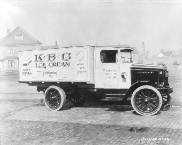 Three-quarter view of passenger side of a KBC Ice Cream delivery truck parked on a cobblestone street. There are houses in the background.