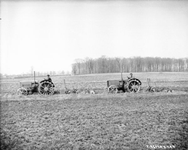 View across field towards two men using tractors to pull plows.