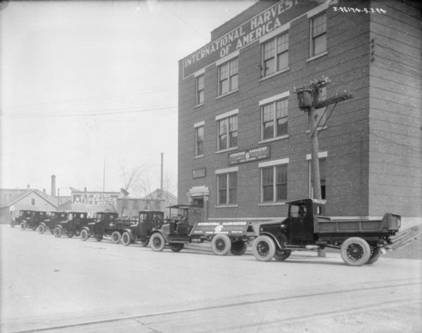 View across street towards a fleet of delivery trucks parked at an angle in a long line in front of an International Harvester of America building. A sign on the back of one of the trucks reads: "McCormick-Deering, Sales, Service Farm Machines."